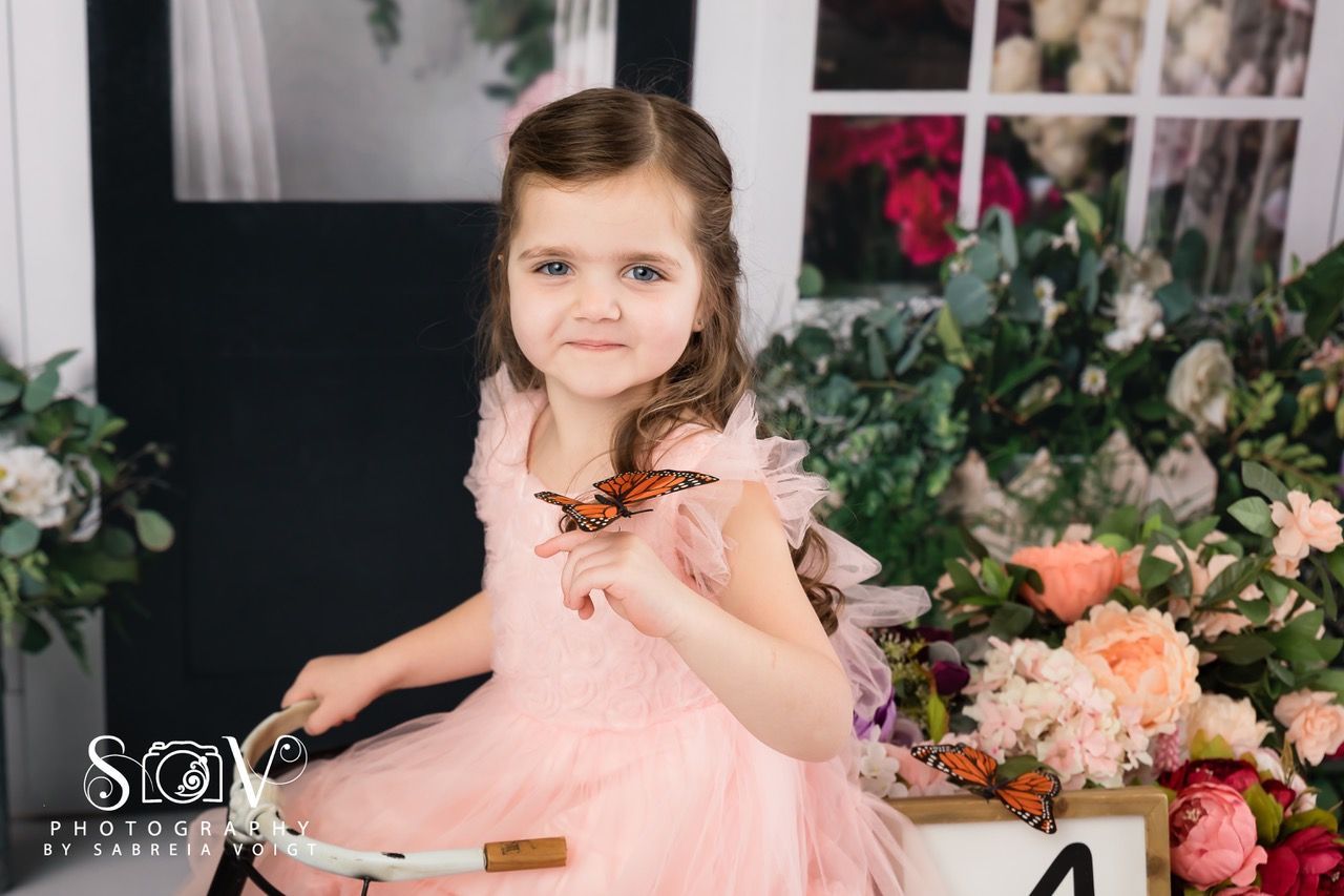 Girl in pink dress with butterflies on her hand, sitting on a bicycle in front of flowers and a decorative door.