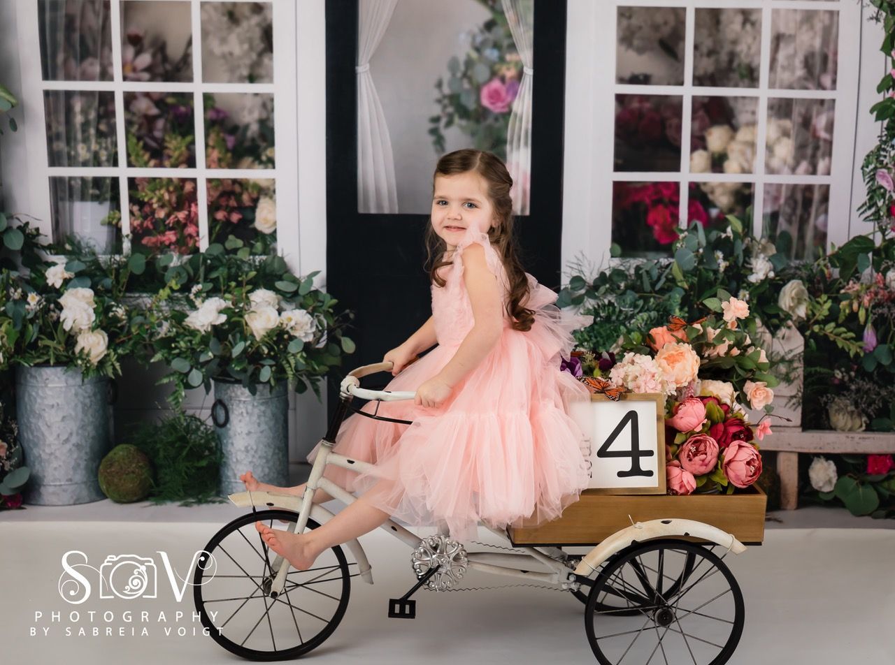 Girl in pink dress sits on tricycle, surrounded by flowers; wooden block reads 