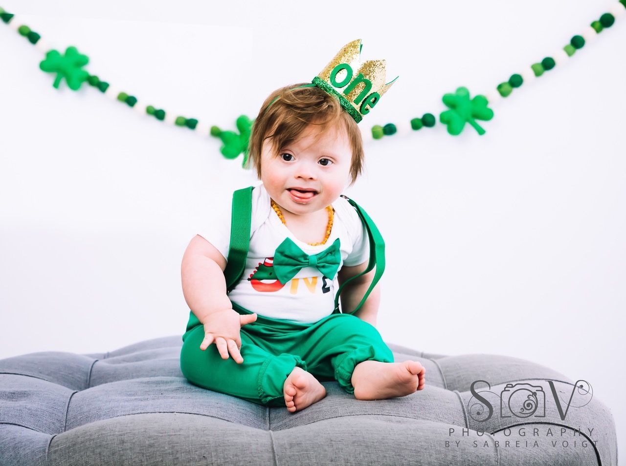 Baby in green outfit and gold crown, sitting on a gray cushion, St. Patrick's Day theme.