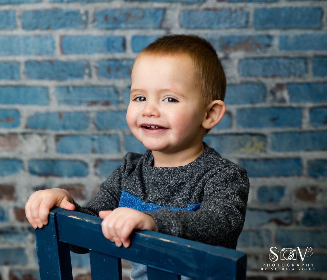 Smiling toddler with brown hair in a gray and blue sweater, holding a blue chair, against a blue brick wall.