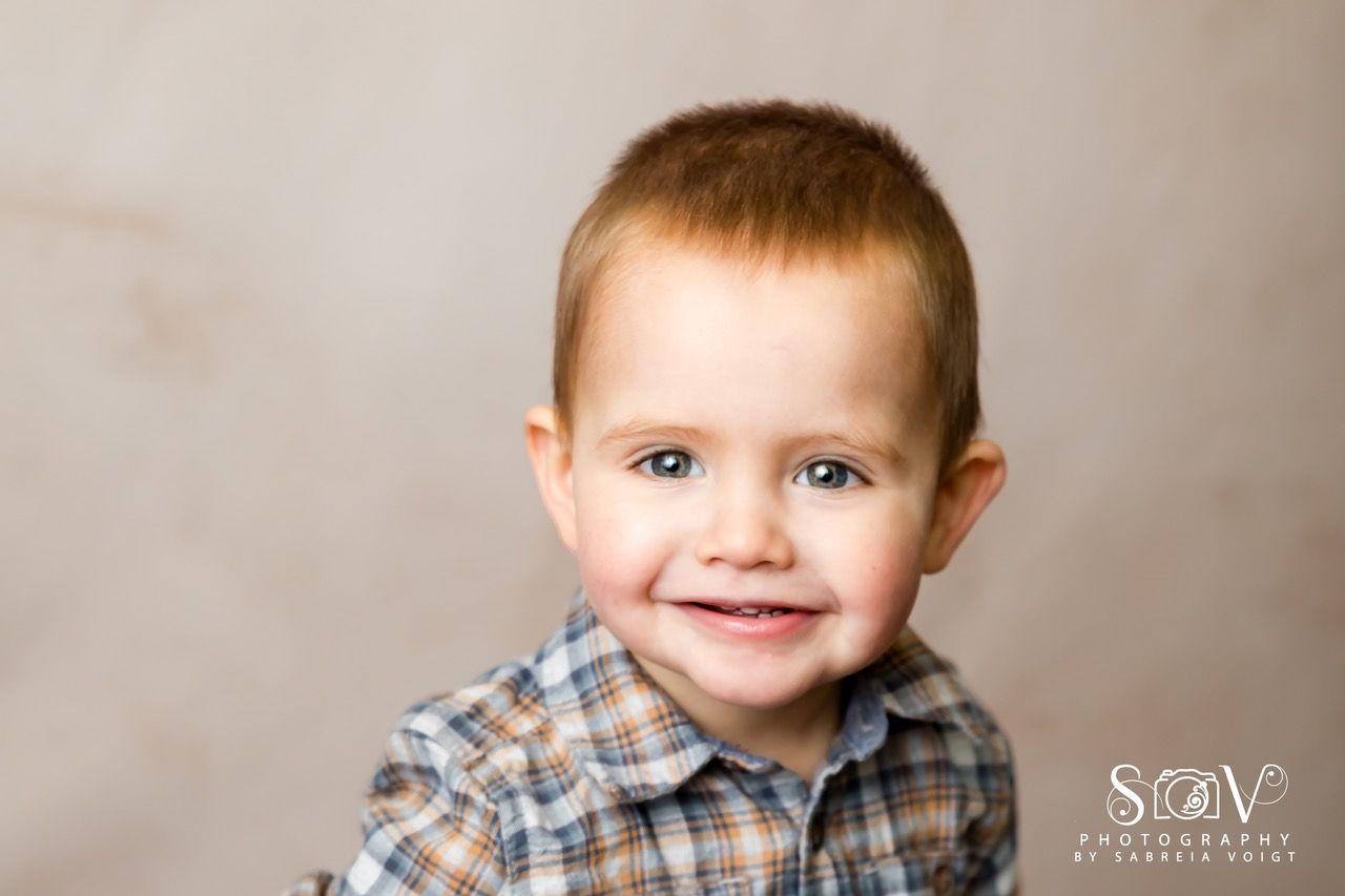 Smiling toddler in plaid shirt, brown hair, against a light brown backdrop.
