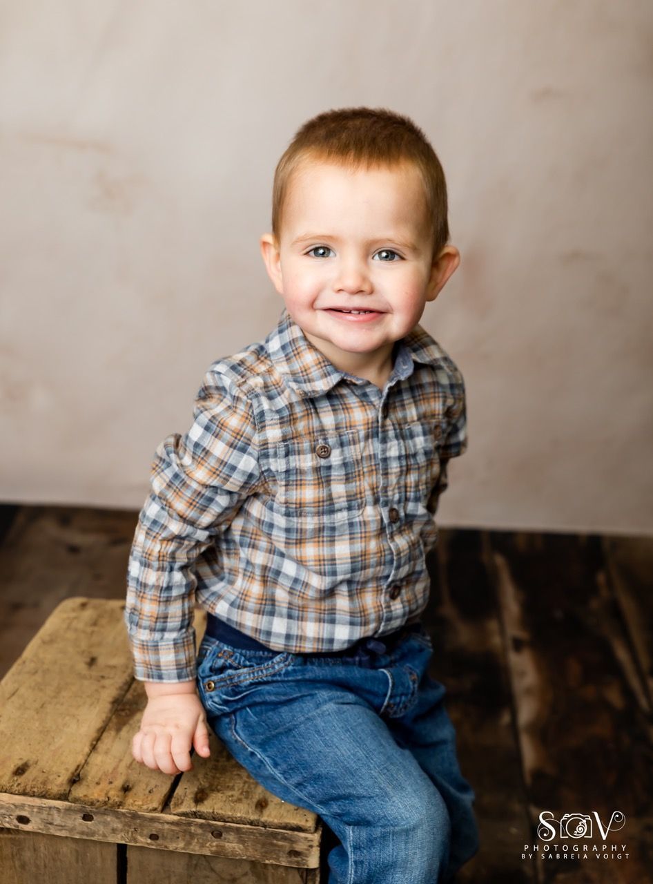 Boy in plaid shirt and jeans, smiling, sitting on a wooden crate.