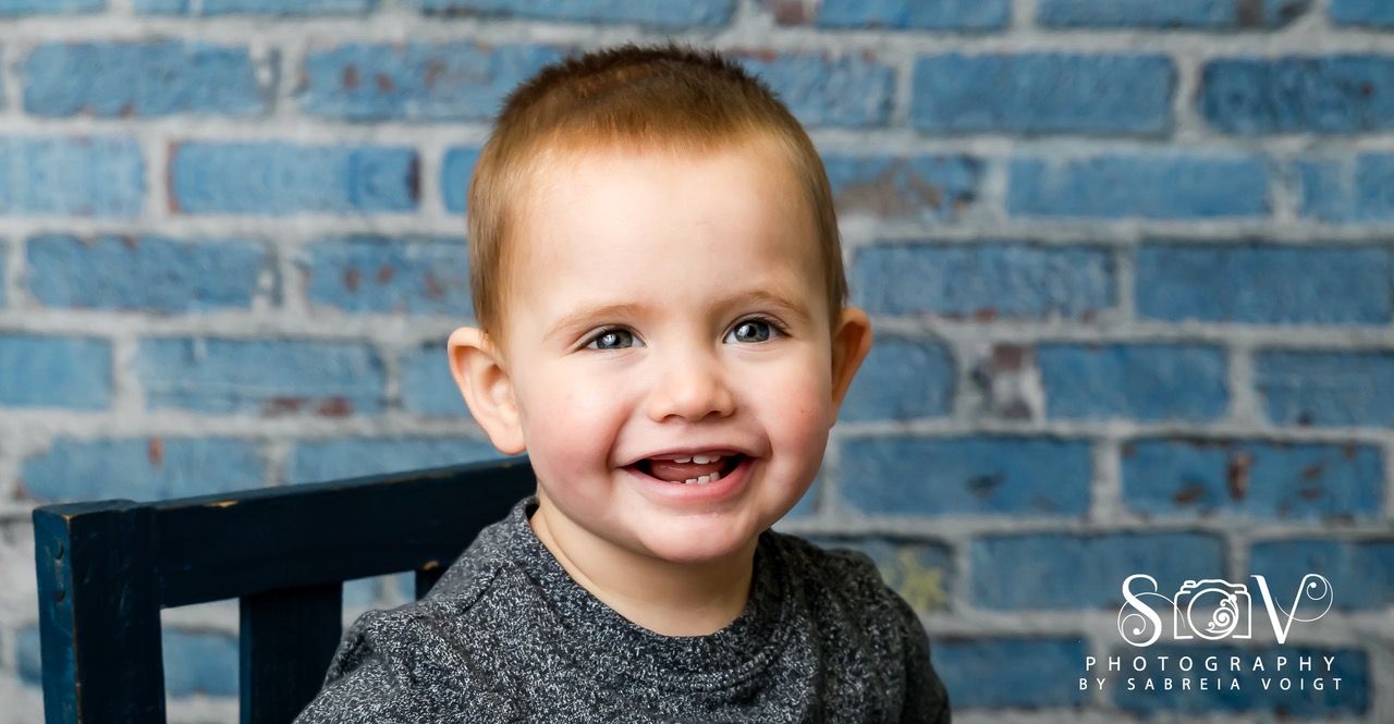 Smiling toddler with reddish-brown hair in front of a blue brick wall.