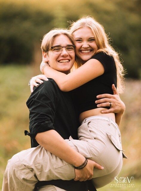 Man holding woman; both smiling, hugging. Woman wearing black top, khaki pants; man wearing glasses, black shirt. Outdoors.