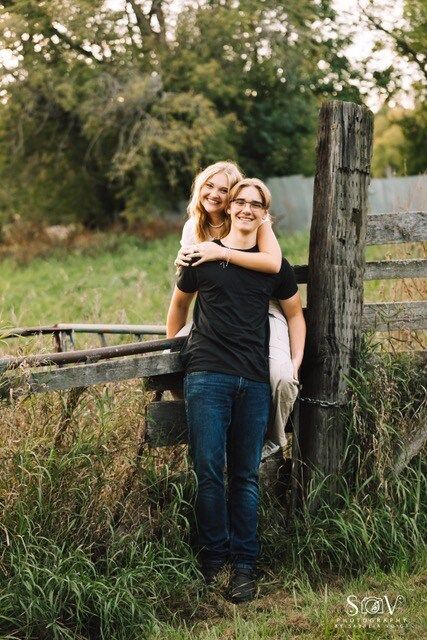 Woman on man's back near a wooden fence. They smile, he wears glasses and black shirt, she has blonde hair.