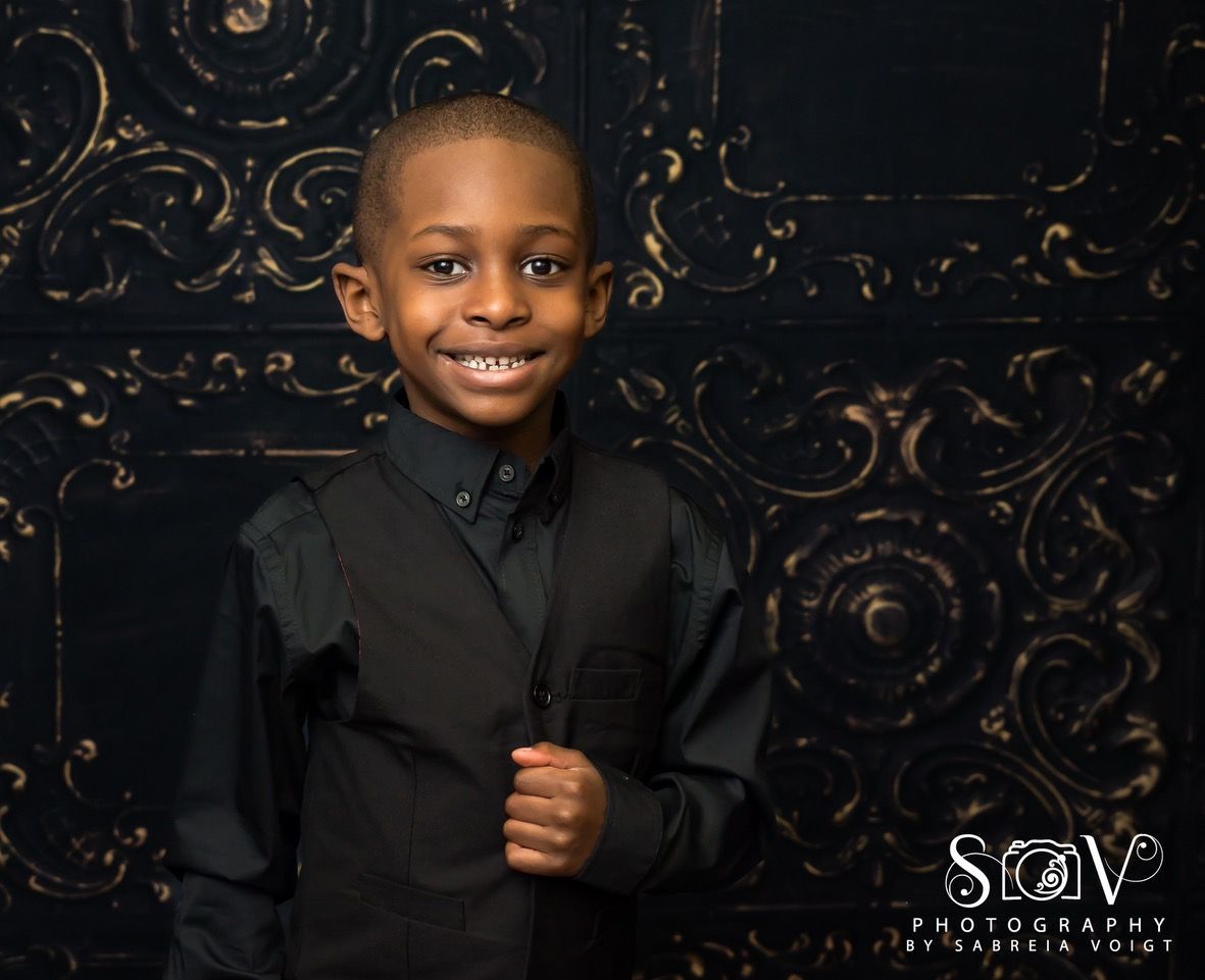 Young boy in black vest and shirt smiles in front of ornate black backdrop.