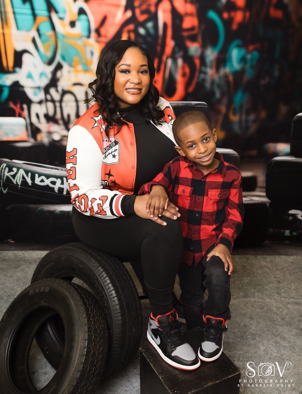 Woman and child sitting, holding hands, in front of graffiti, wearing jacket, black shirt/pants, and plaid shirt/pants.
