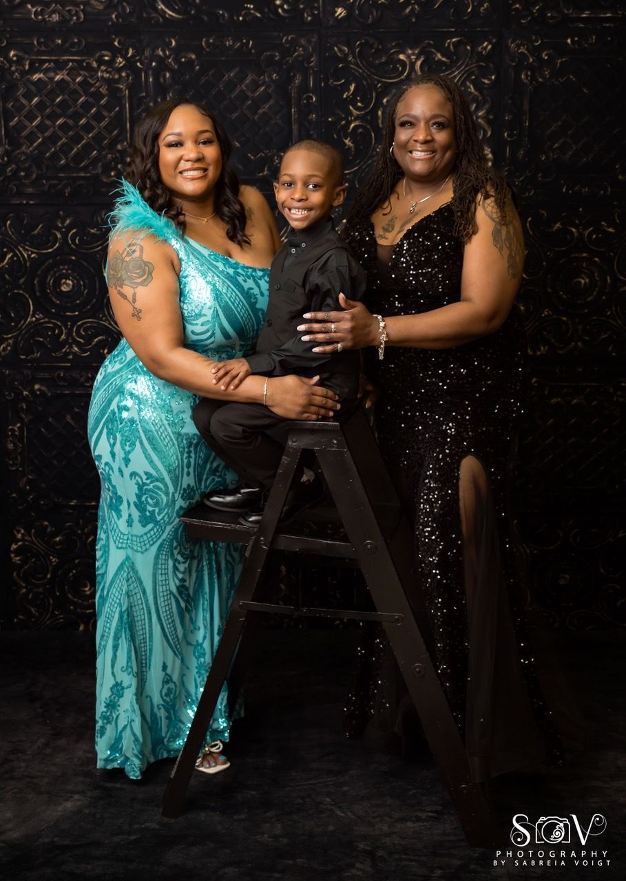 Woman in teal gown, boy in black suit, and woman in black gown pose for photo. Dark background.