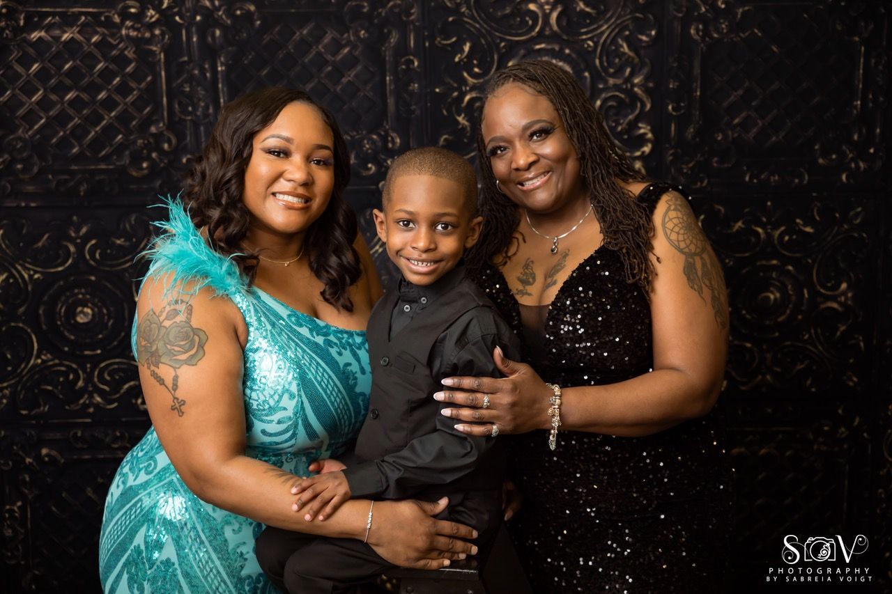 Woman in turquoise dress with a young boy and a woman in a black sequined dress posing in front of a dark backdrop.