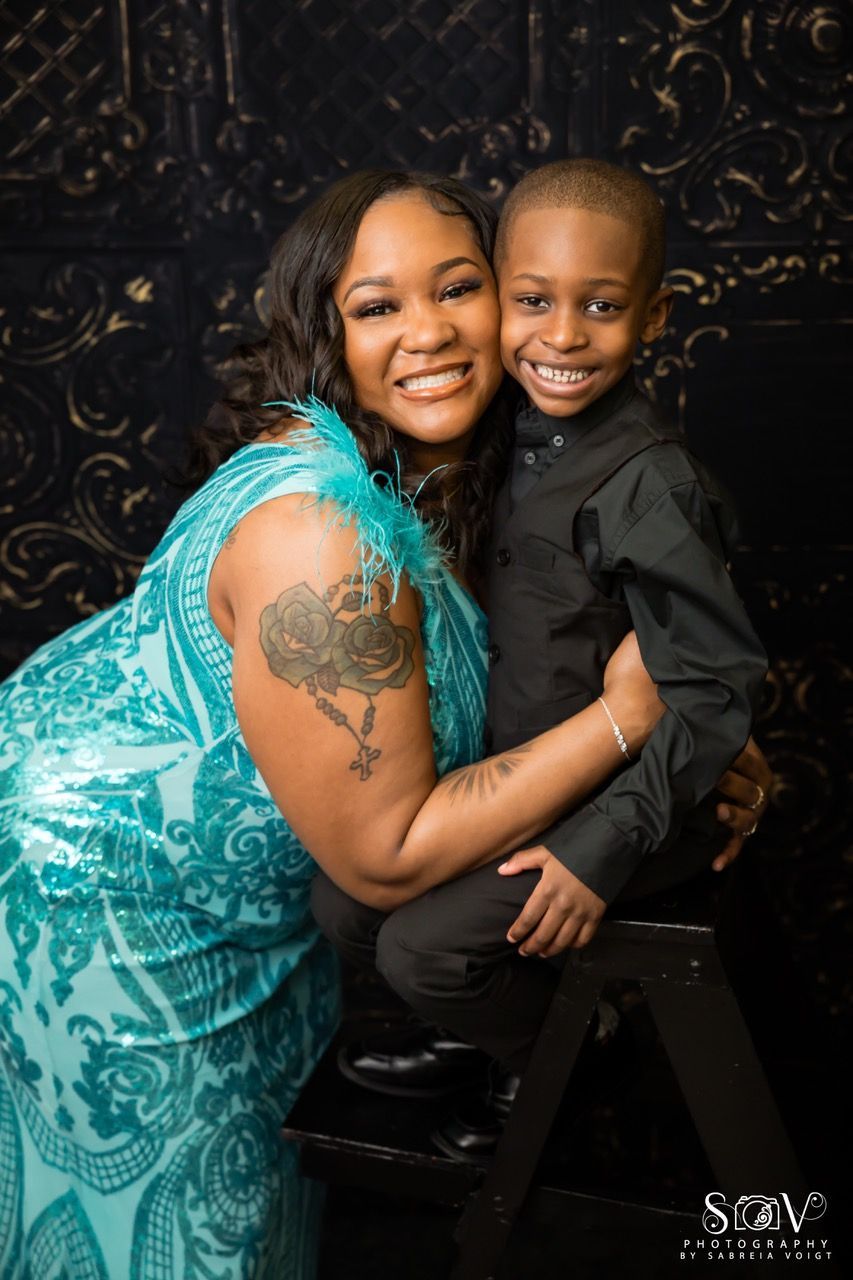 Woman in teal dress hugs a young child in black clothing. Both smile in front of a decorative background.