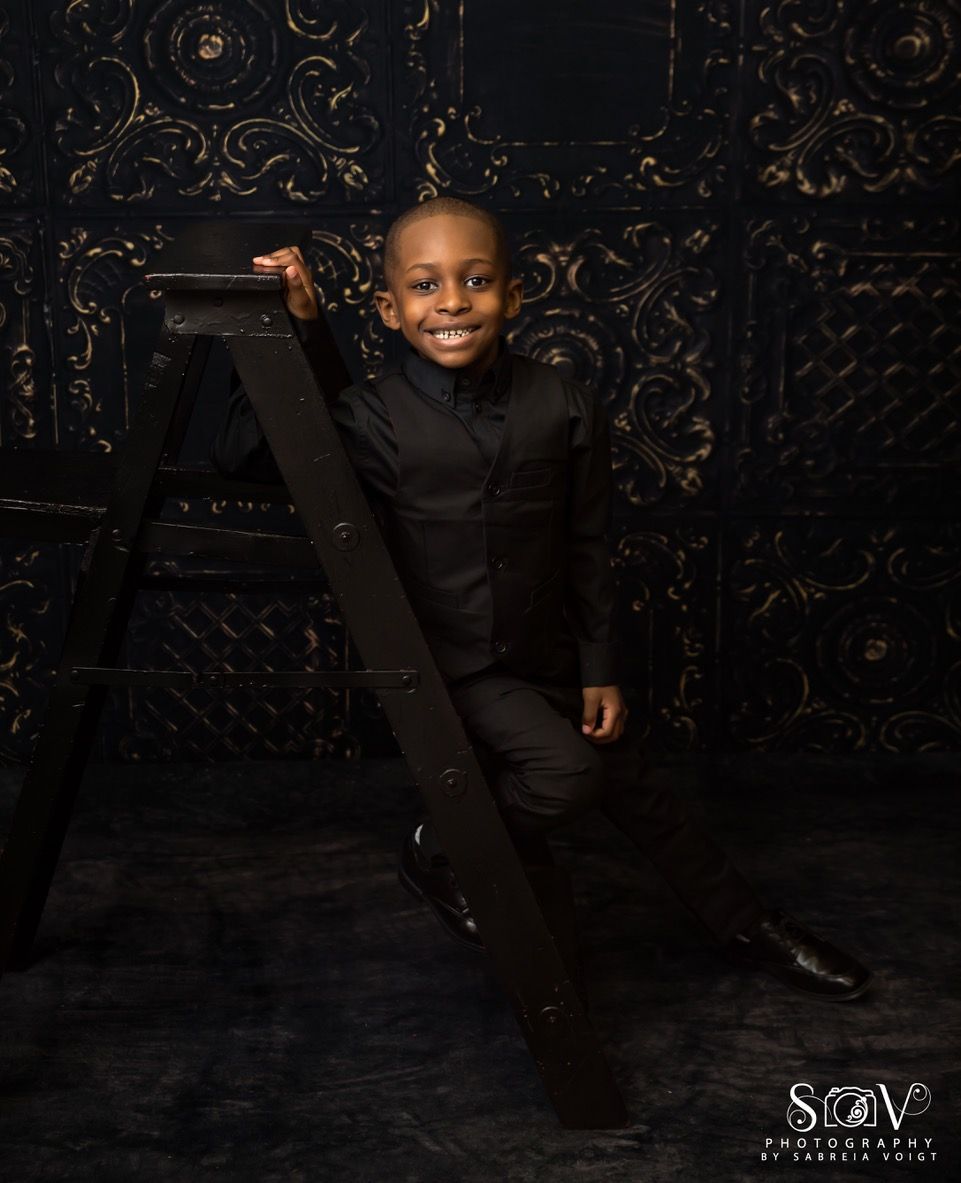 Boy in black suit smiles, leaning against a black ladder. Dark, ornate background.