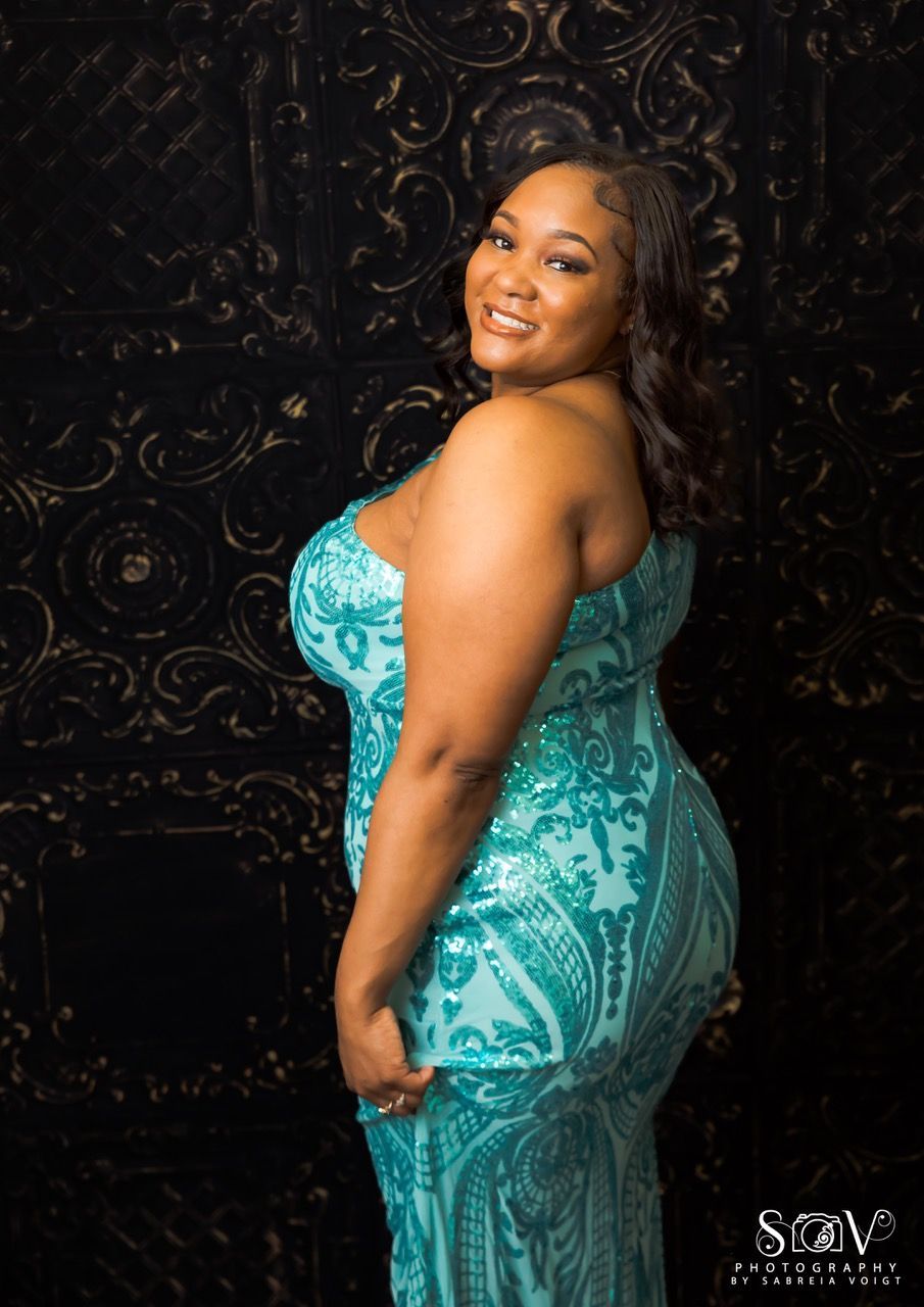 Woman in a teal, sequined, one-shoulder gown smiles. Stands against a decorative black backdrop.