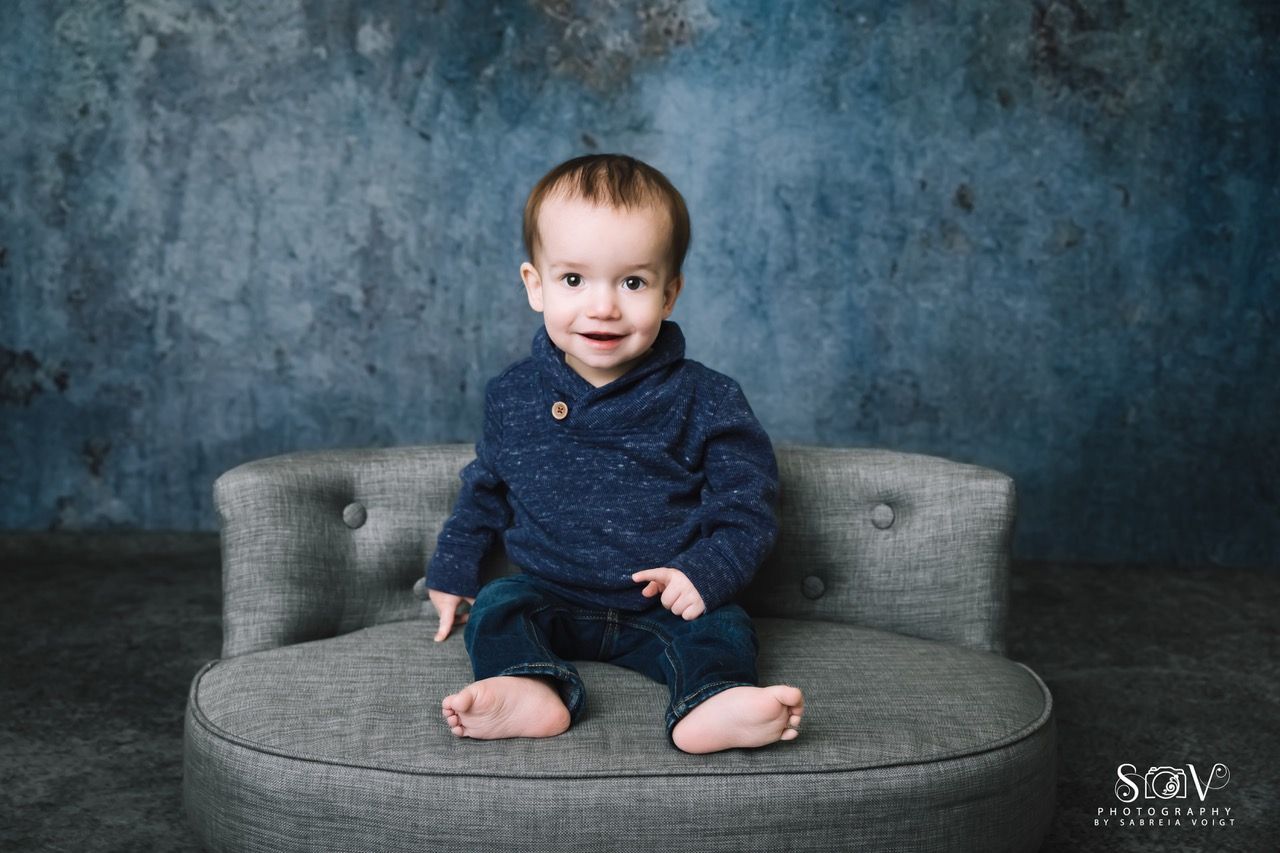 Baby seated on a gray ottoman, wearing blue sweater and jeans; blue textured backdrop.