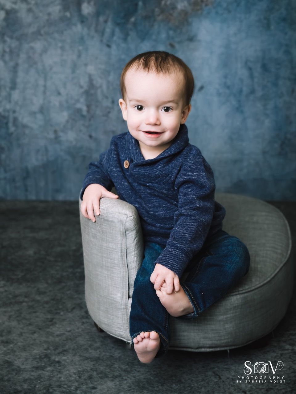 A young child sitting in a gray round chair against a textured blue backdrop, smiling.