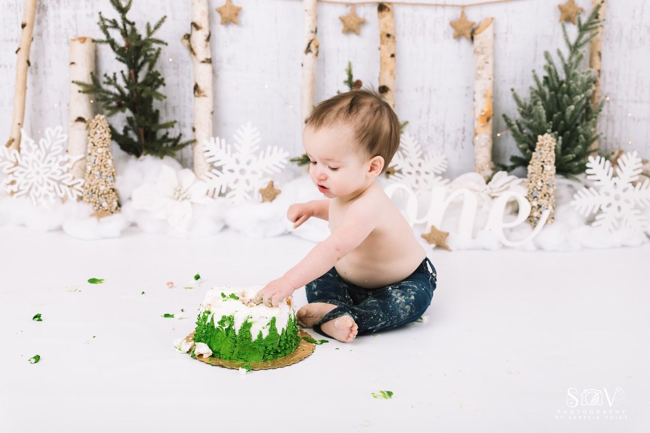 A baby sits in front of a winter-themed backdrop, playing with a green cake.