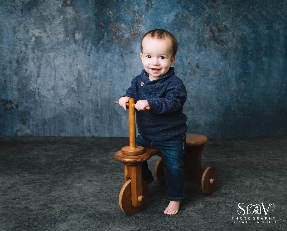 Toddler on a wooden tricycle, smiling, wearing blue sweater and jeans. Against a blue textured backdrop.