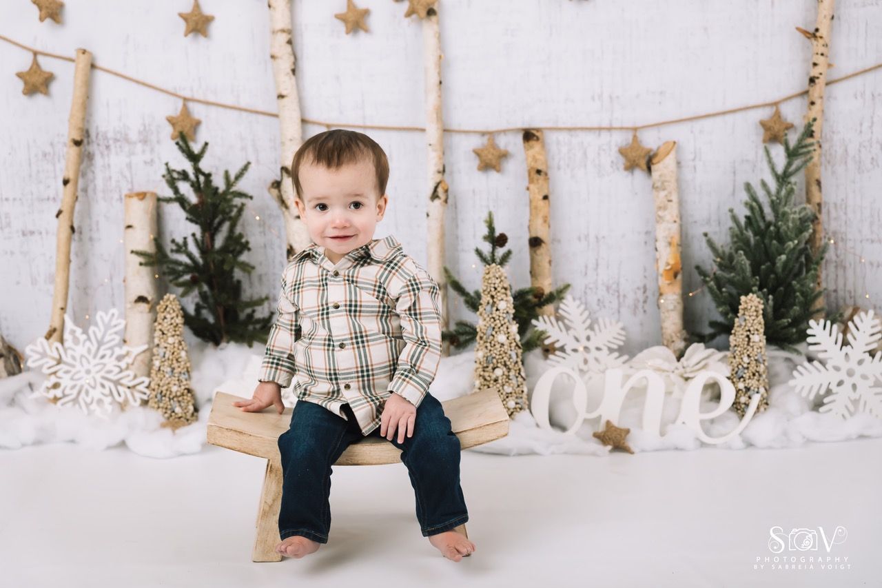 A toddler sits on a wooden bench, smiling at the camera. Festive winter backdrop with decorations.