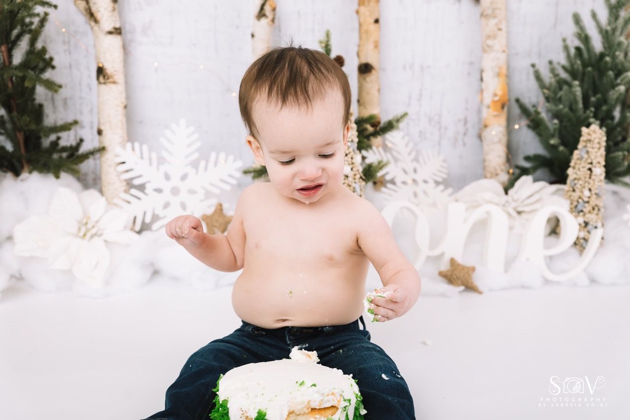 Bare-chested toddler sits atop a cake, smearing frosting. Winter-themed backdrop with 