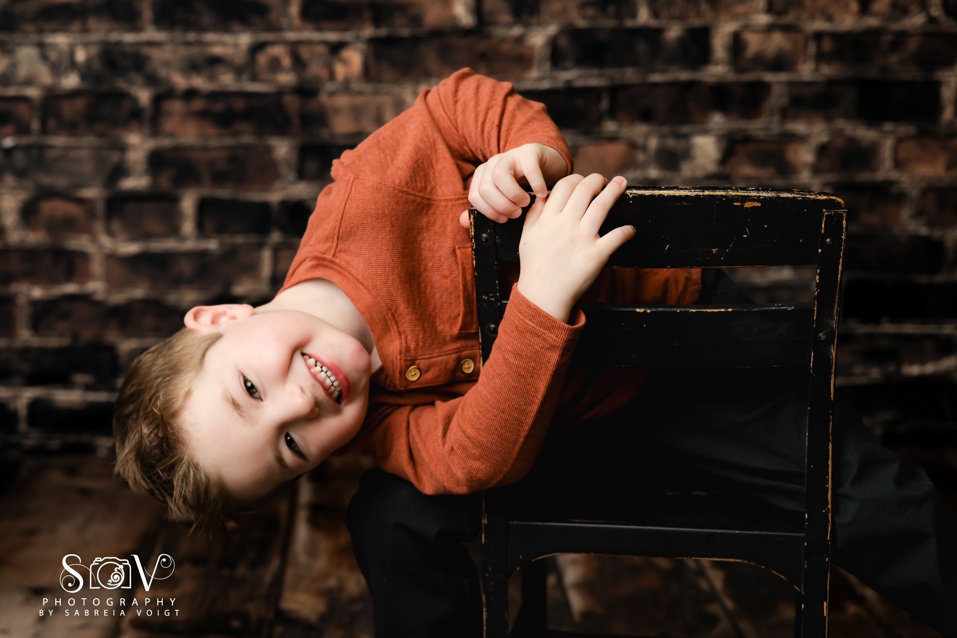 Smiling child in orange shirt leans over a black chair against a brick wall.