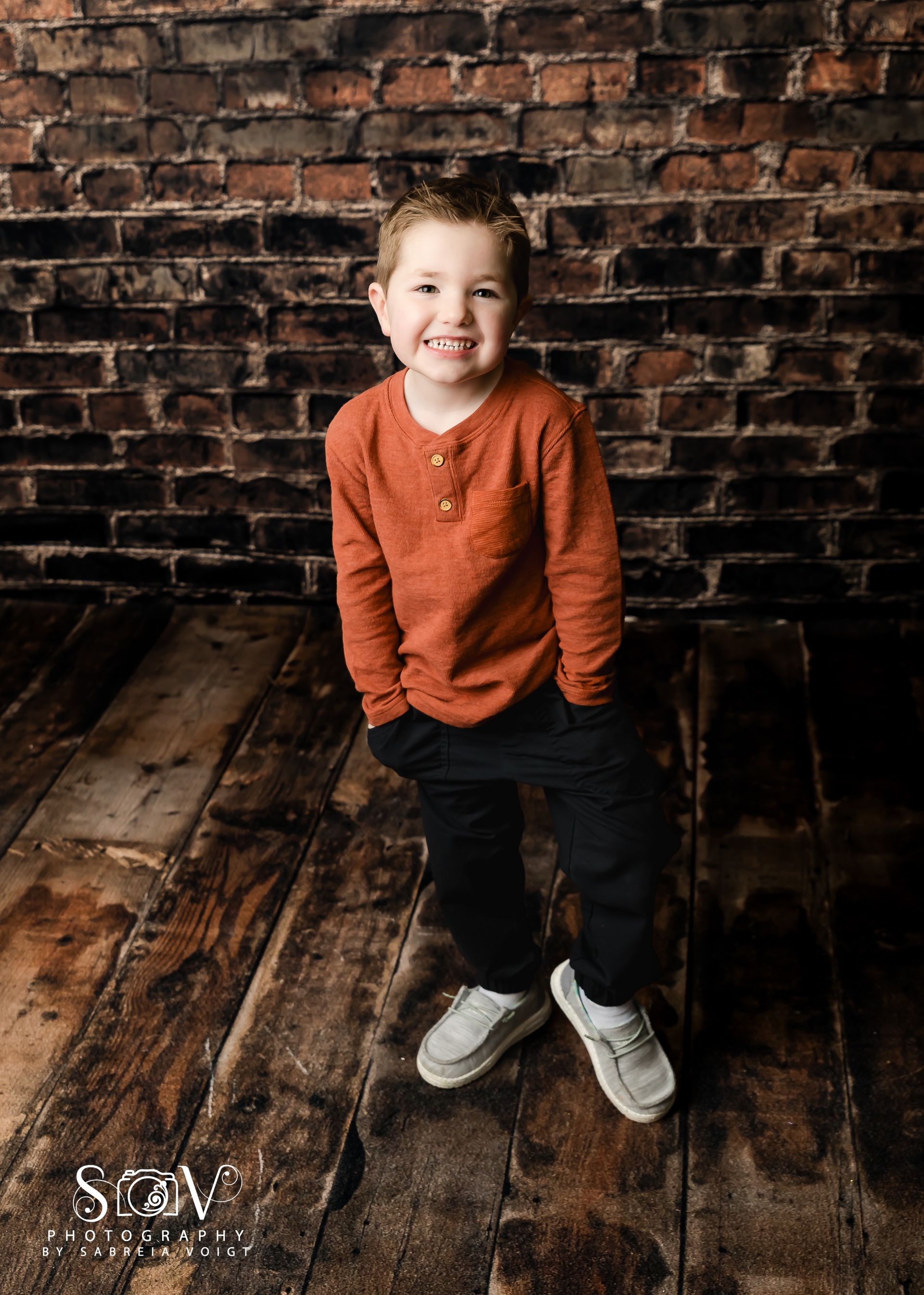 Boy smiling in orange shirt and dark pants, hands in pockets, against a brick wall and wooden floor.