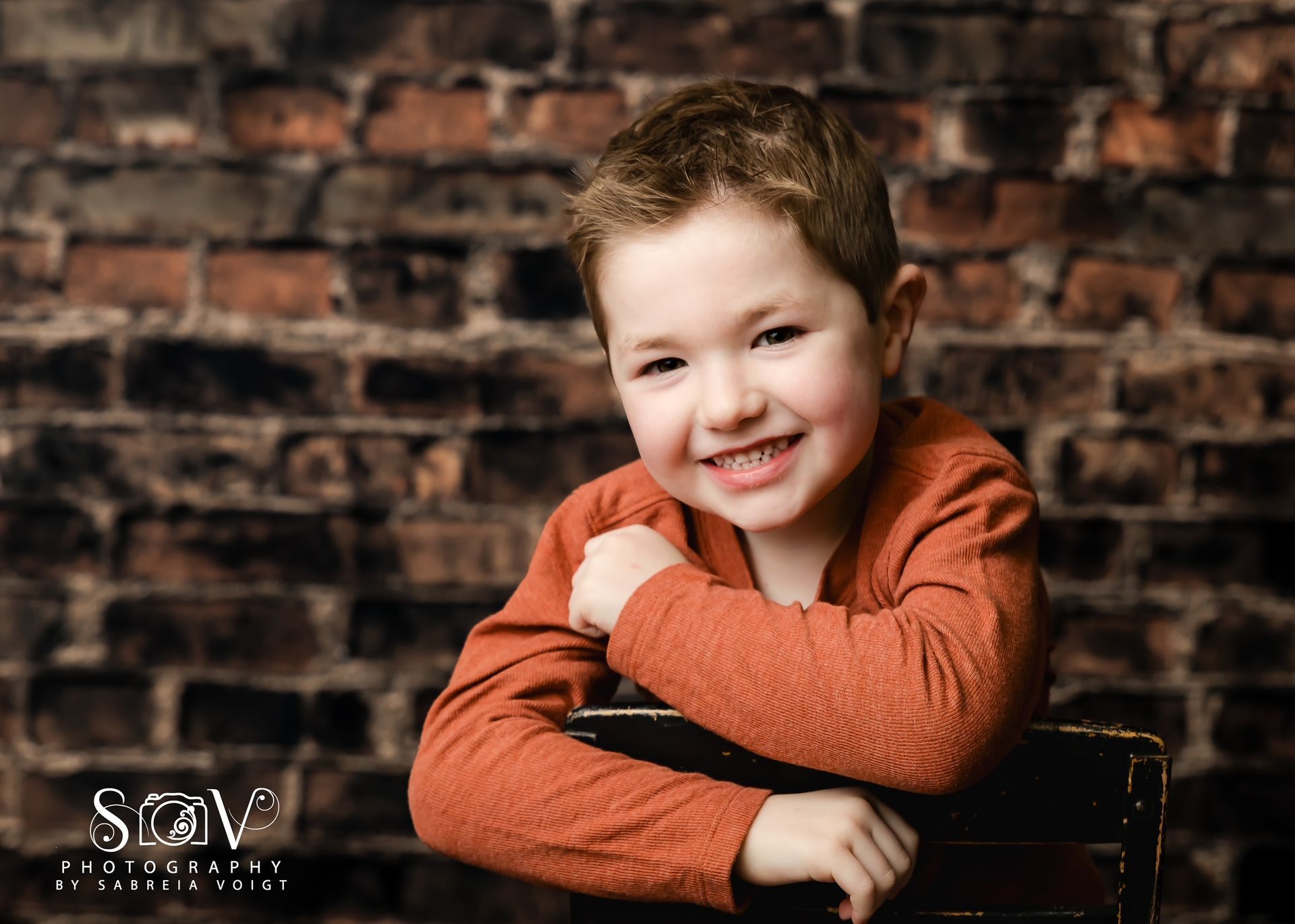 Young child smiling, resting arms on a chair, against a brick wall. Wearing a rust-colored shirt.