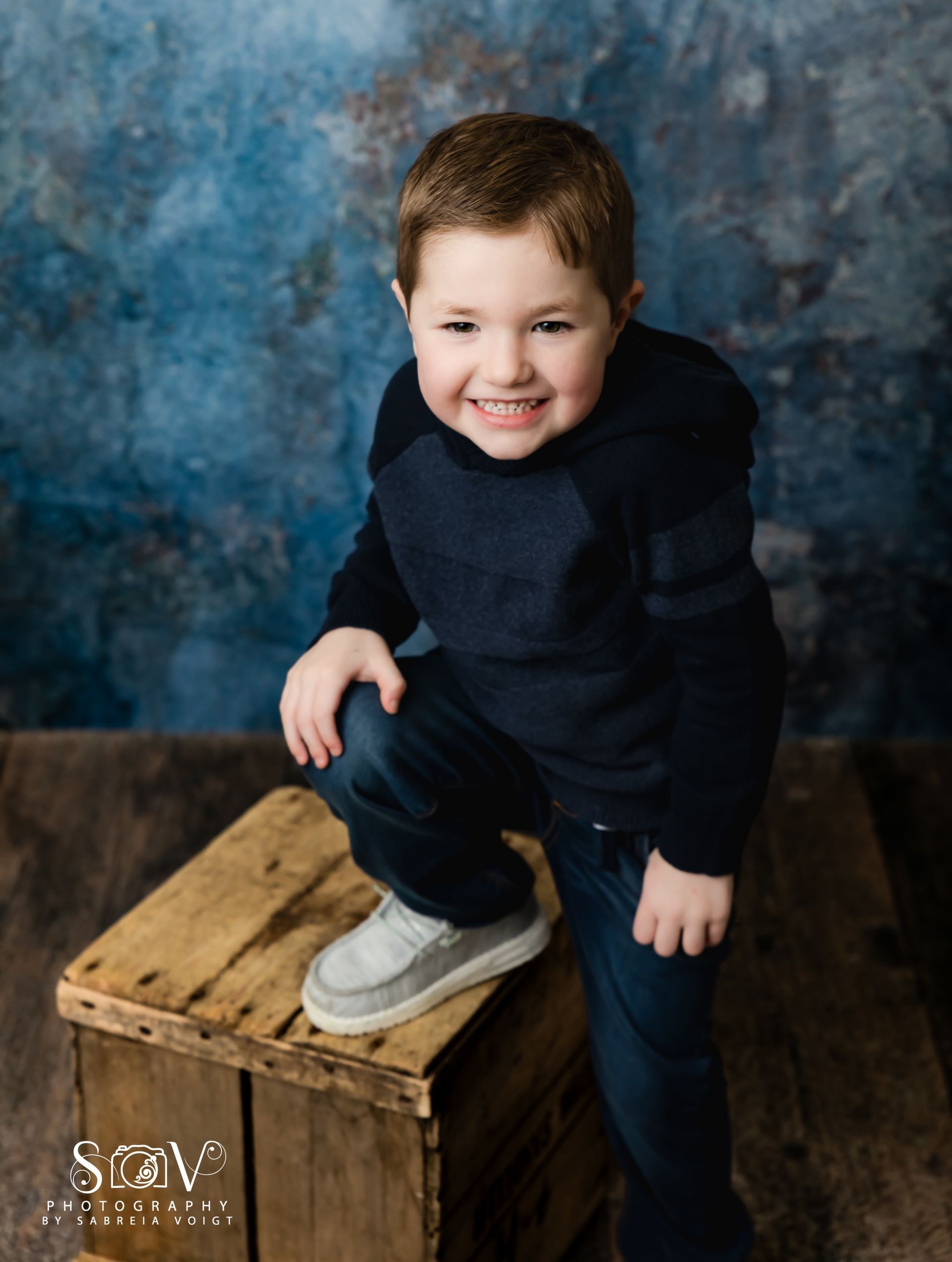 Boy smiles, posed on a wooden box, wearing a navy blue shirt and jeans; blue background.