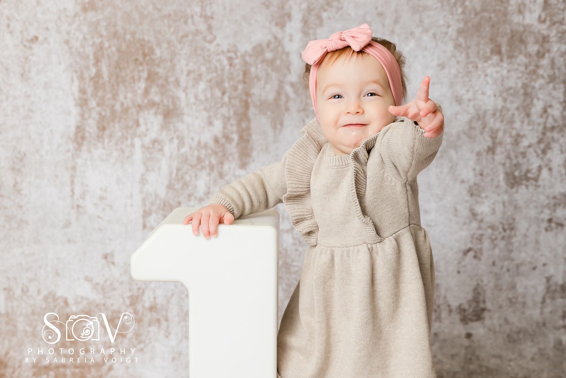 Girl in light brown outfit with pink headband giving a thumbs-up, standing beside a large number 
