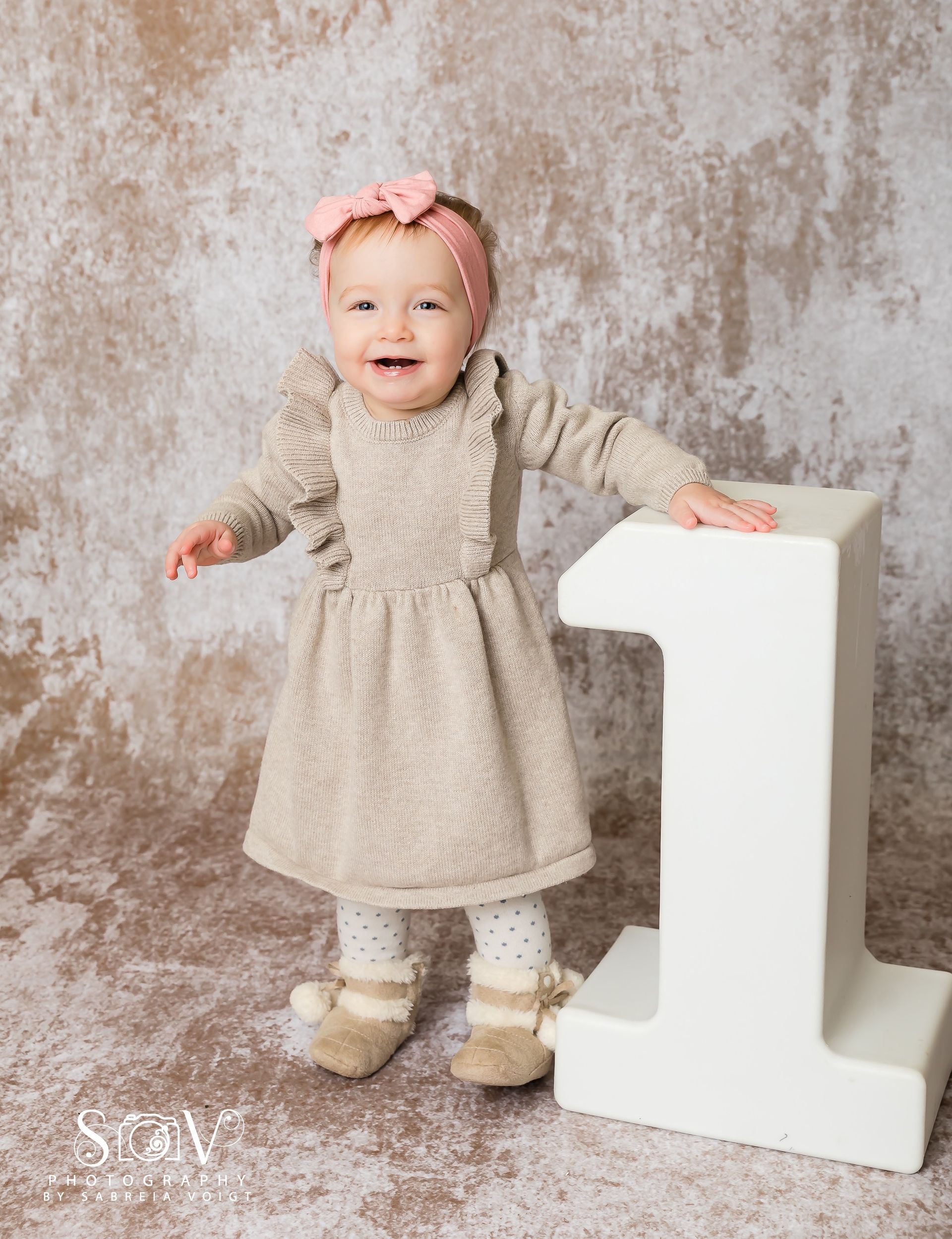 Smiling toddler wearing a beige dress and a pink bow headband leans on a white number one.