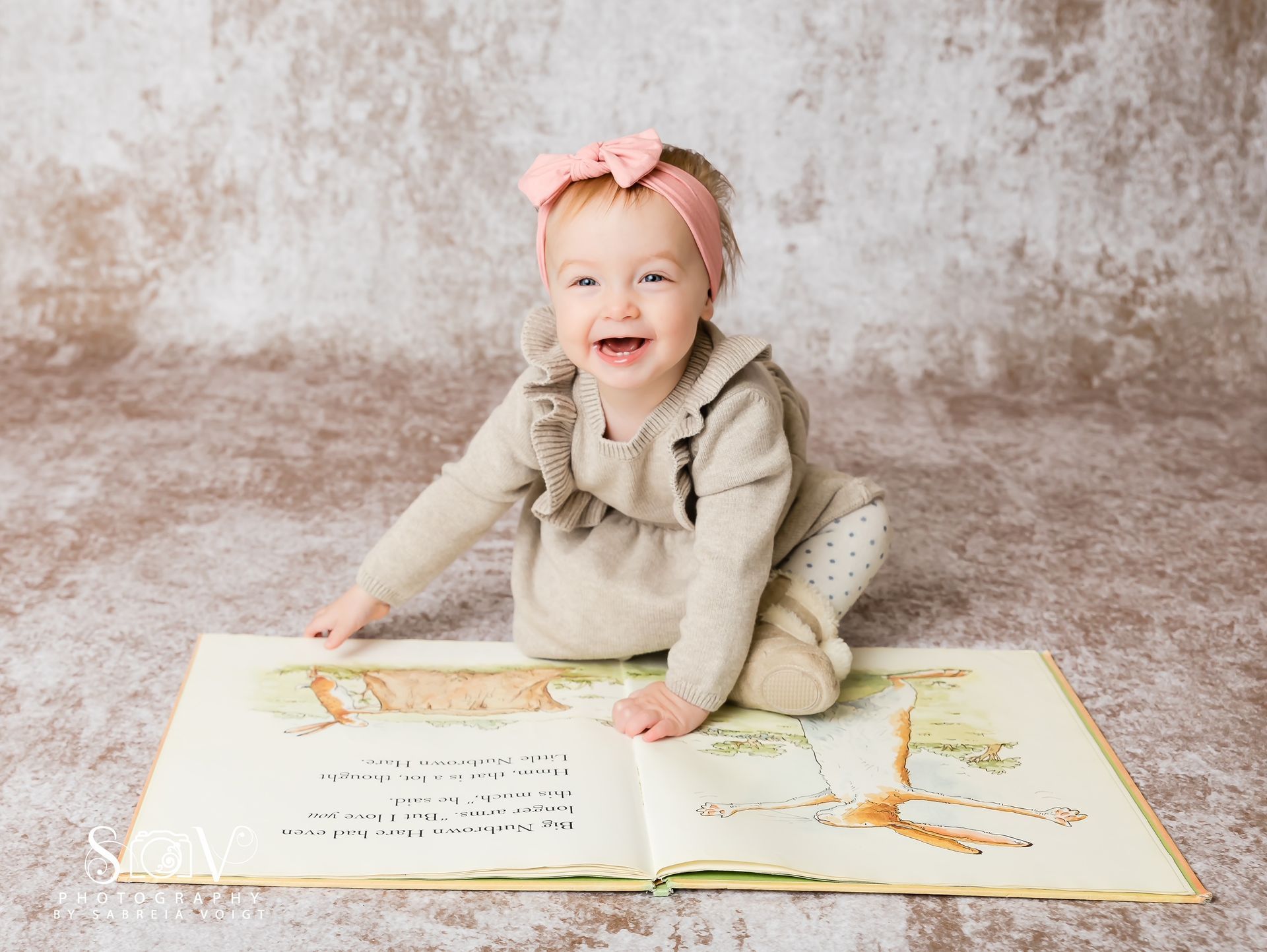 Smiling baby with pink headband, reading an open book on a mottled brown surface.
