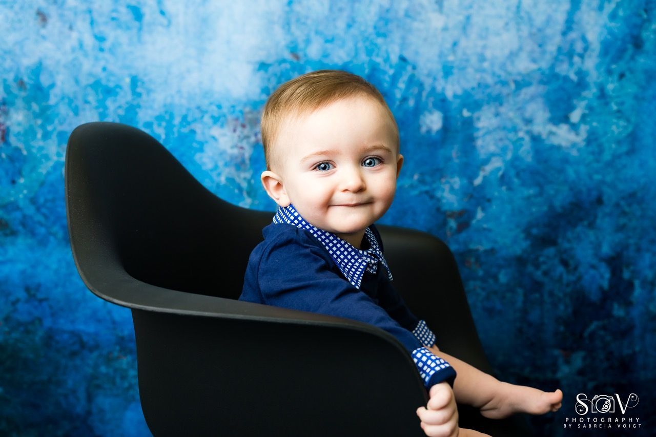 Smiling baby in a black chair against a blue abstract background, wearing a blue shirt and bowtie.