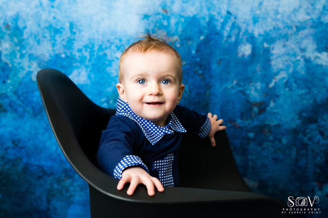 Baby with blue eyes smiles in a black chair against a blue, textured backdrop.