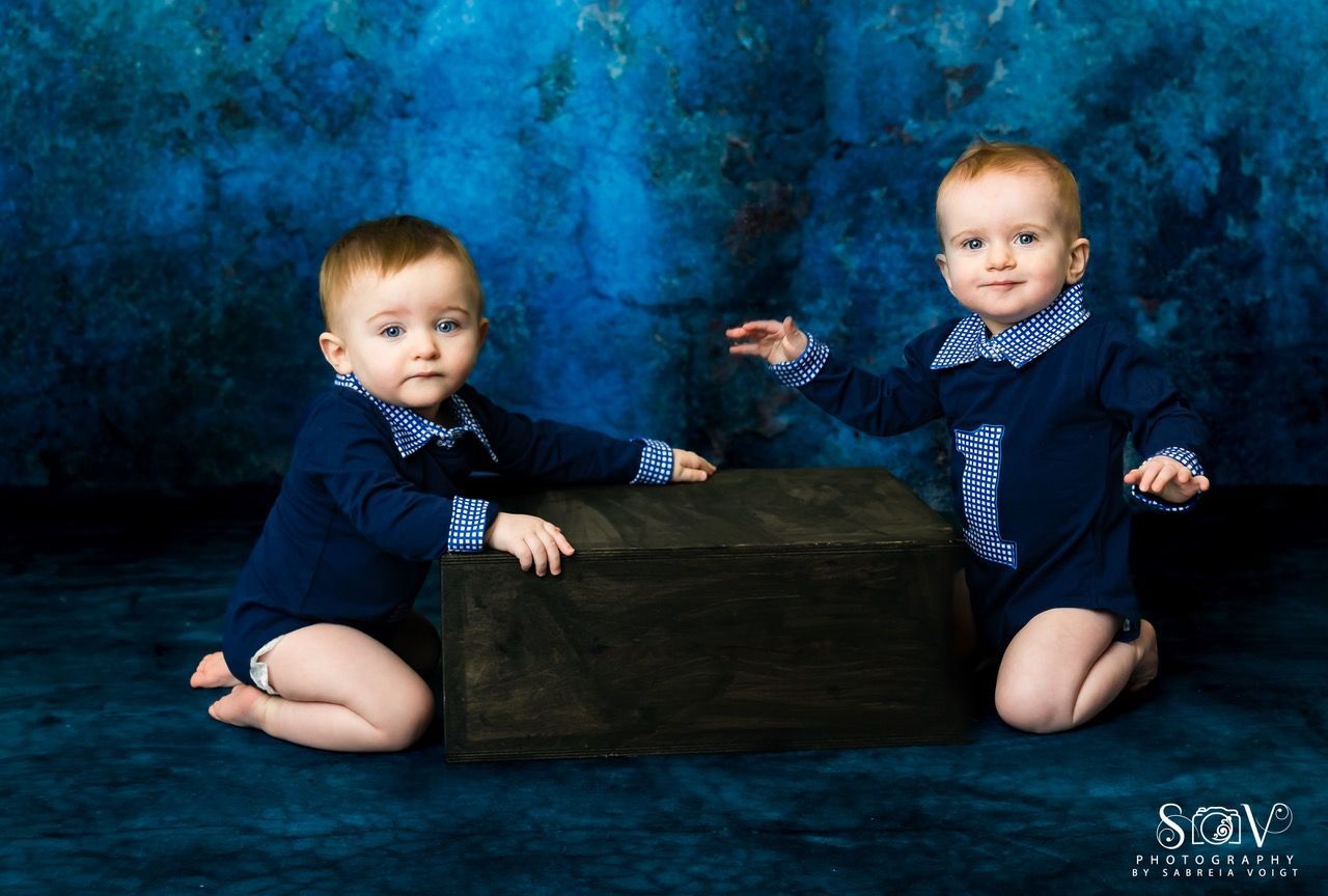 Two babies in blue outfits on their knees, looking at the camera, with a dark box between them.