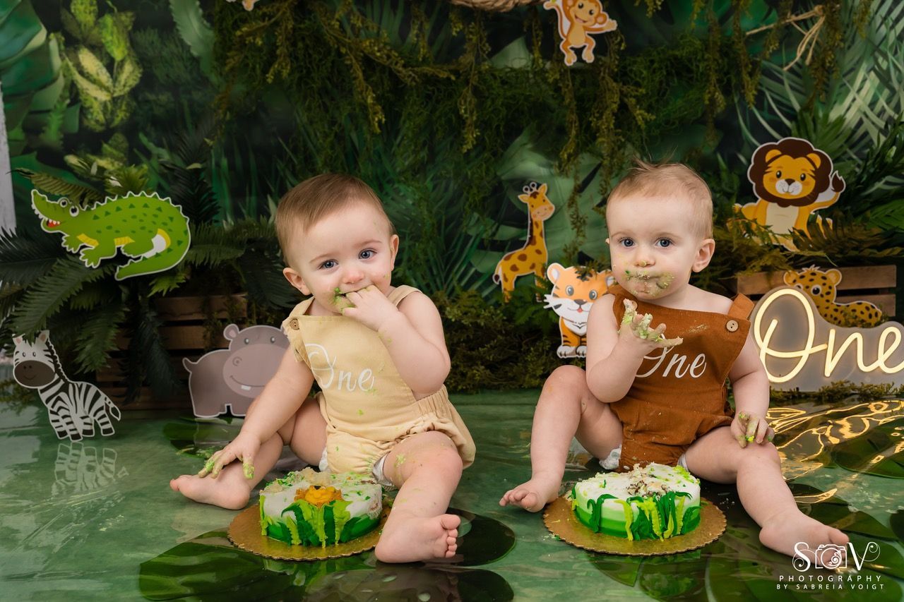 Two babies in jungle setting with cake, eating cake, decorated with jungle animals.