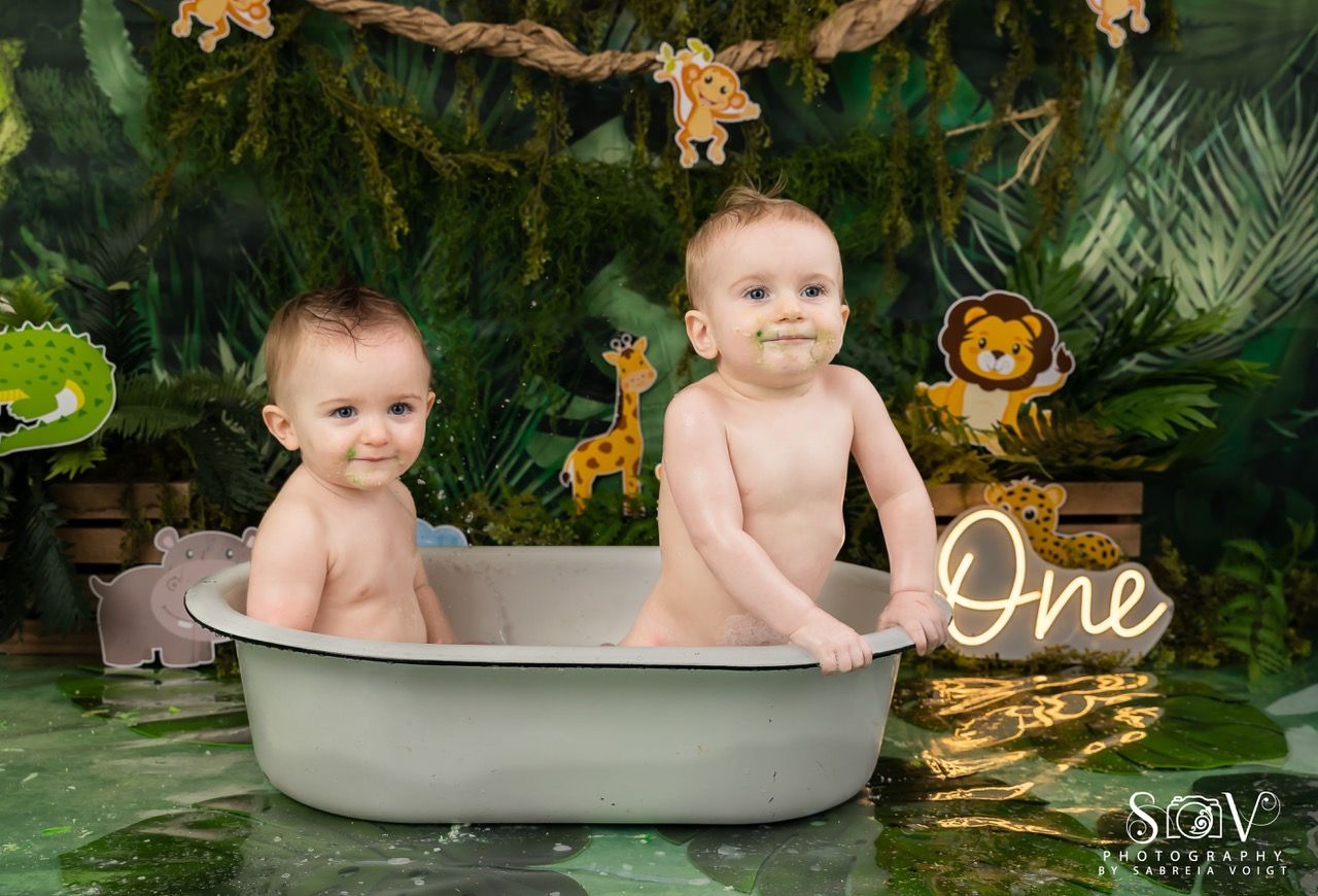 Two babies in a tub, jungle-themed. One with food on their face, smiles. Surrounded by jungle decor and 