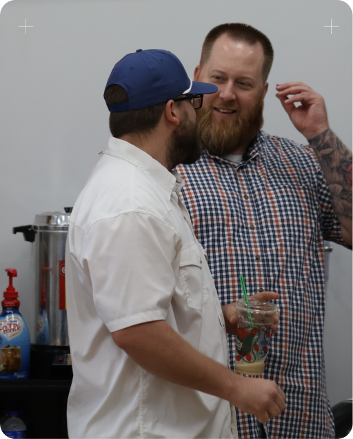 Two men chatting at an indoor event, one in a white shirt and blue cap holding a drink.