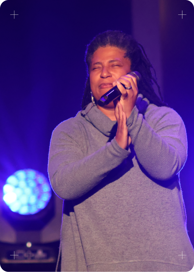 Person singing into a microphone on a purple-lit stage, wearing a gray sweater.
