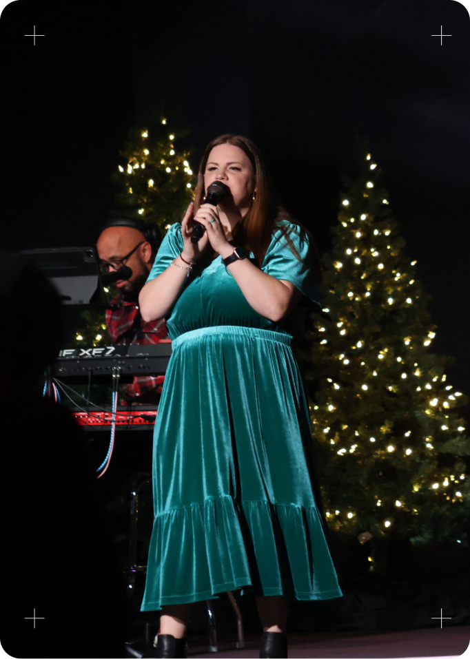 Singer in a teal dress performing onstage with microphone, lit Christmas trees behind her.