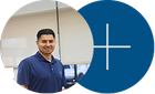 Man in a navy polo standing beside a blue circle with a white plus sign in an office setting