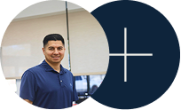 Man in a navy polo standing indoors beside a dark blue circle with a white plus sign