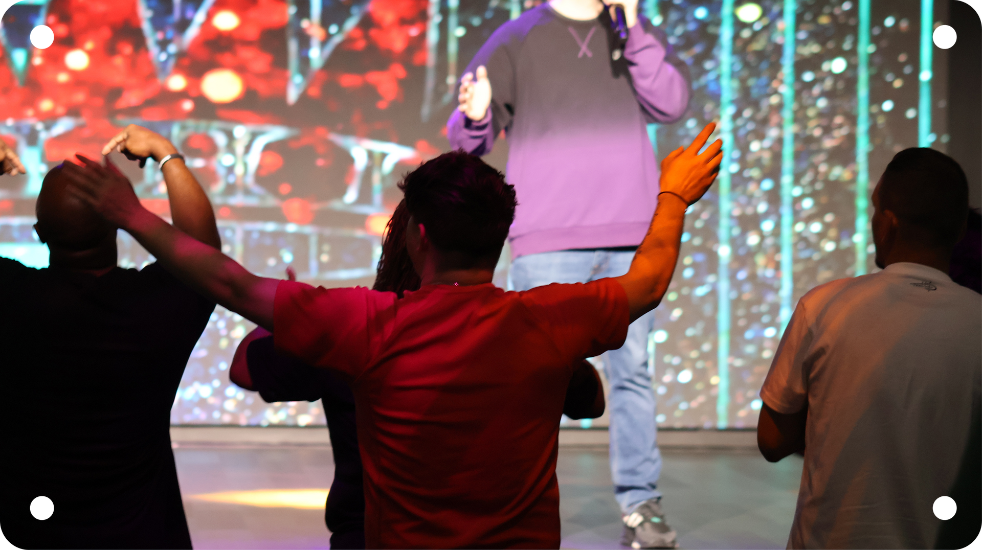 Audience member in a red shirt raises both arms toward a performer on a lit stage with a colorful backdrop