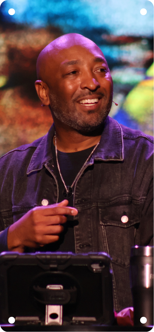 Smiling man in a dark jacket on stage, holding a microphone with colorful lights behind him