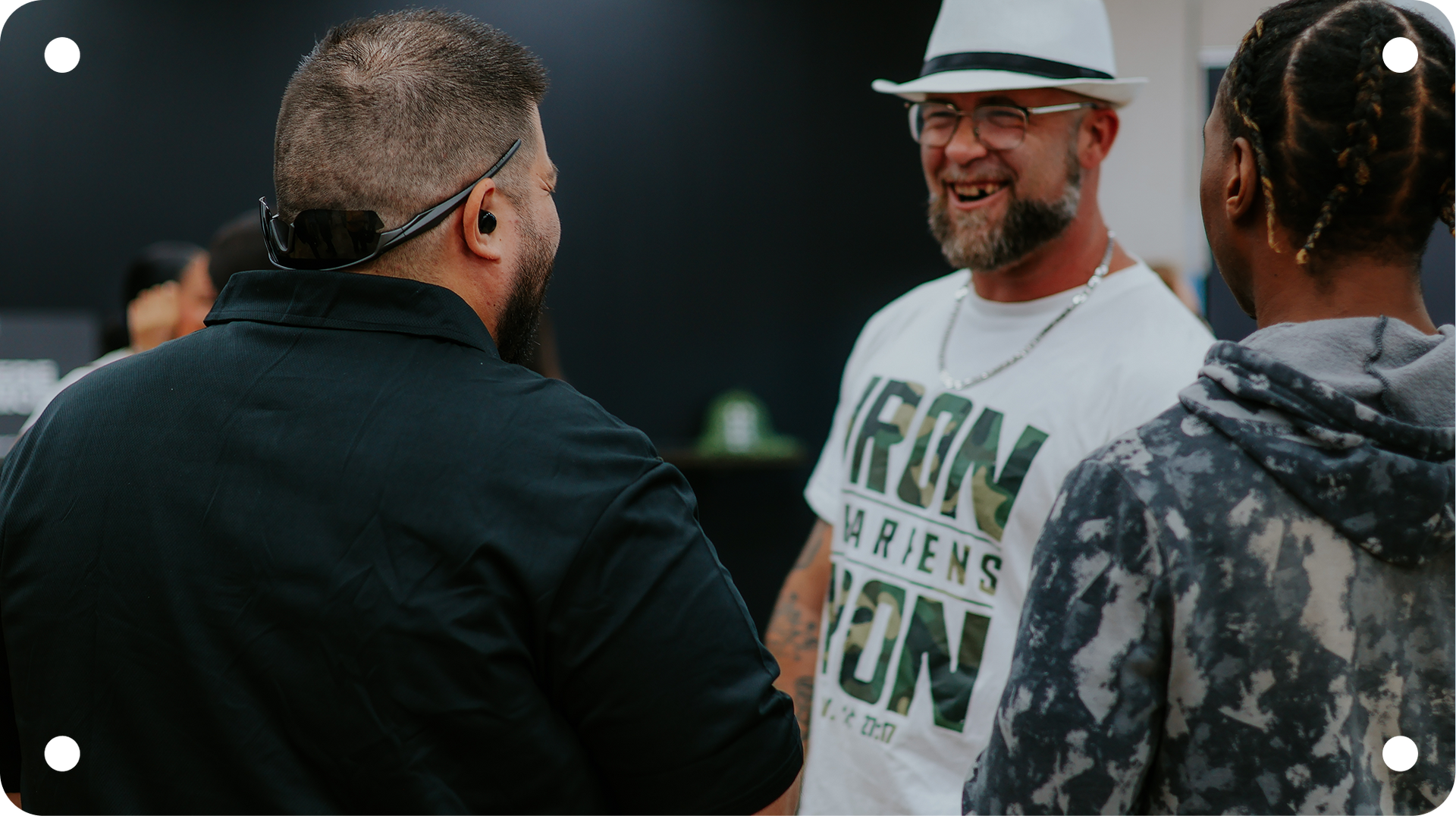 Three people talking outdoors, one smiling in a white hat and green shirt with “IRON MEN RUN” text