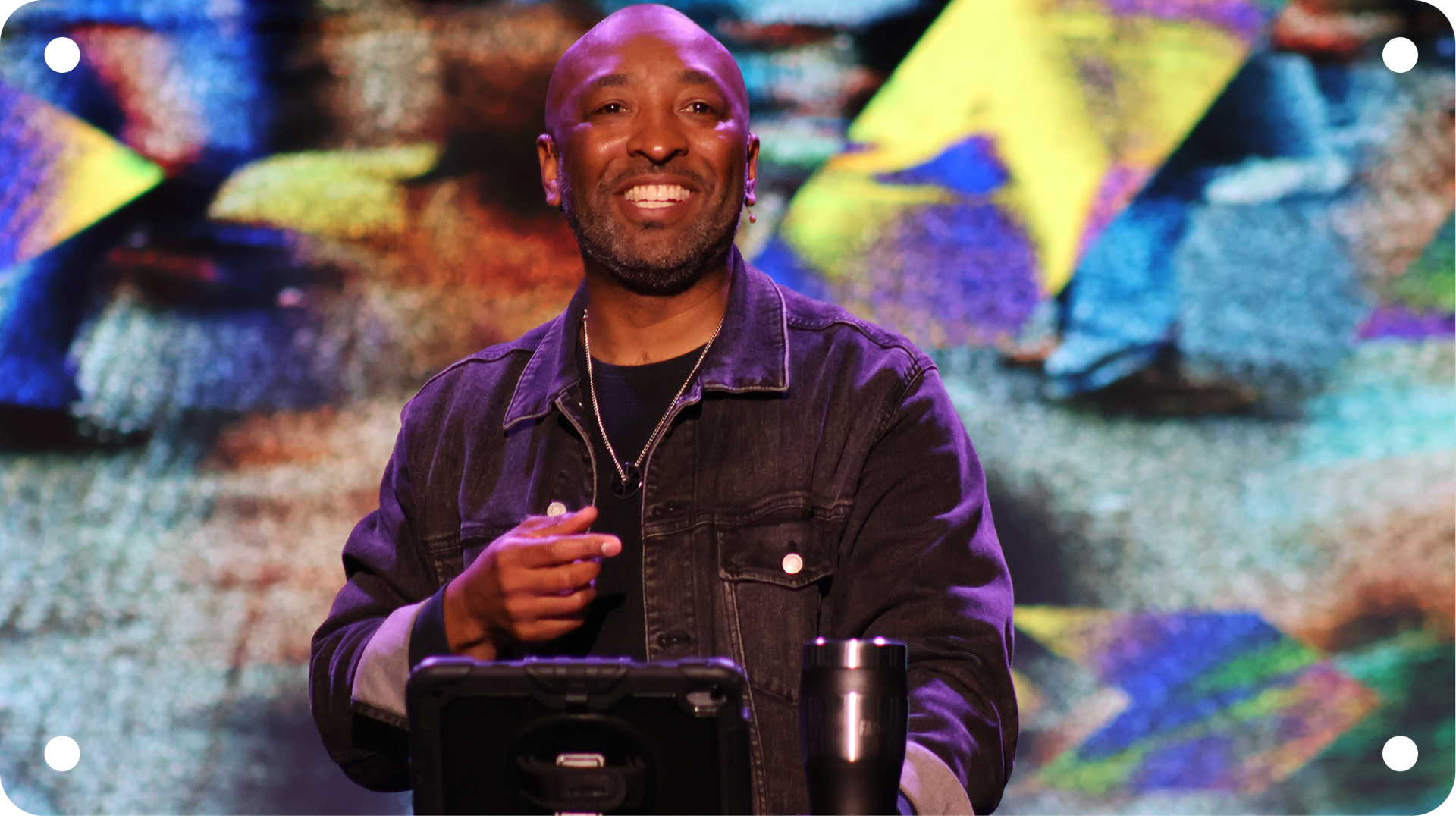 Smiling speaker at a podium on stage, holding a microphone with colorful abstract lights behind him