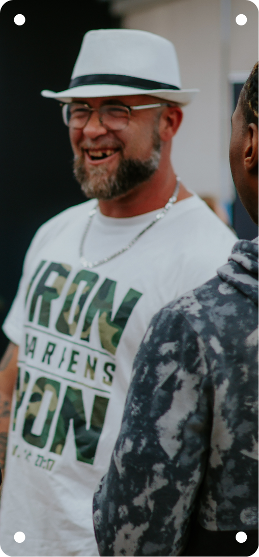 Smiling man in white hat and shirt with green text, standing outdoors beside another person