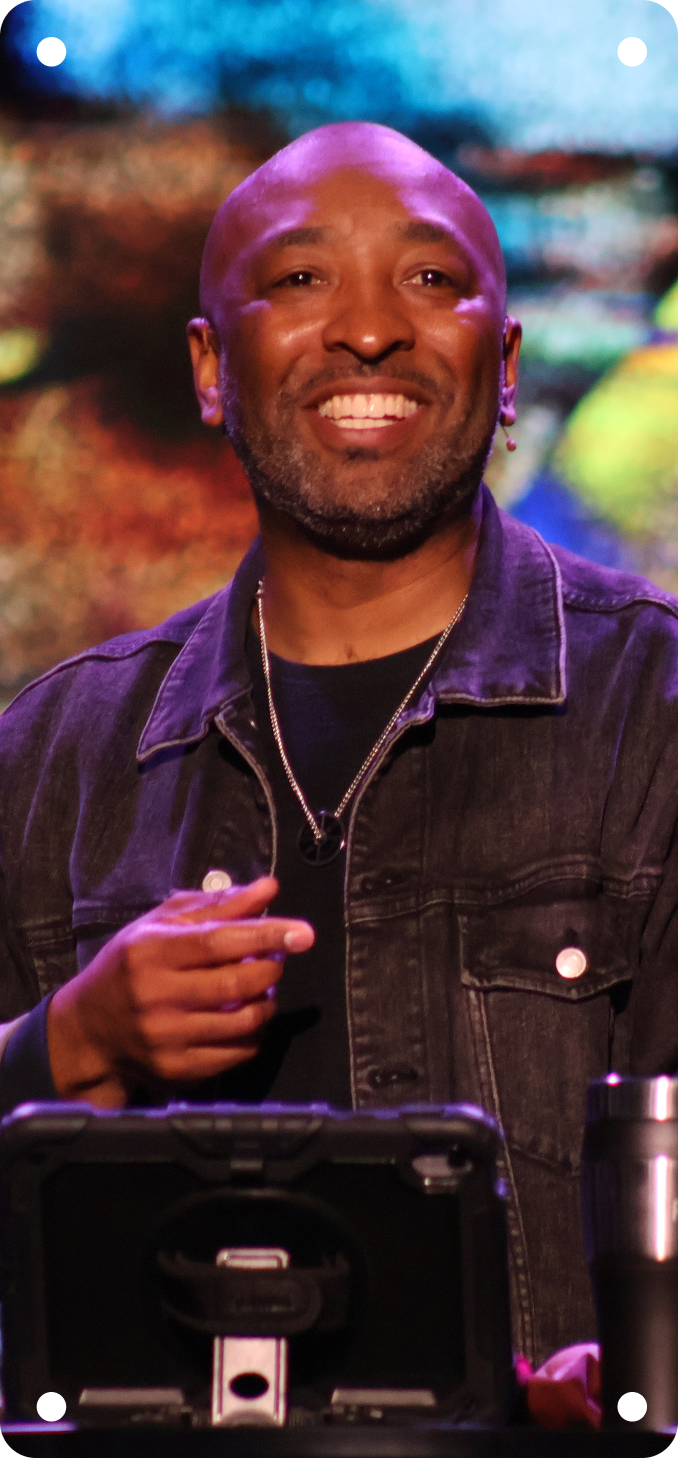 Smiling man in a denim jacket speaking on stage under colorful lights