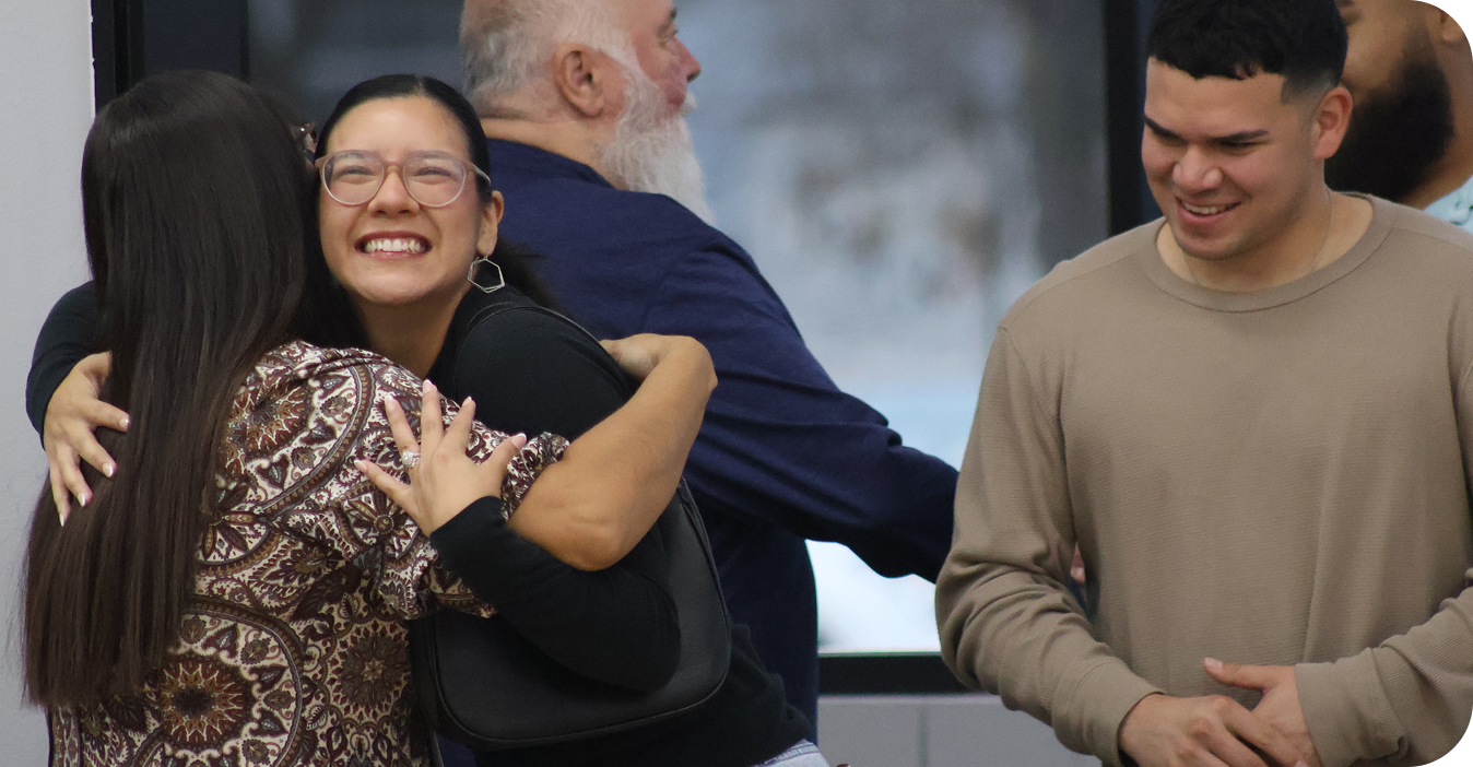 Three people smiling and hugging indoors, one in a black shirt and cream cardigan
