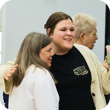 Three people smiling and hugging indoors, one in a black shirt and cream cardigan