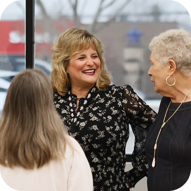 Three women chatting indoors, smiling near a window in a bright room.