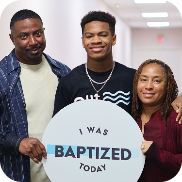Three people smiling in a hallway holding a sign that says, “I was baptized today.”