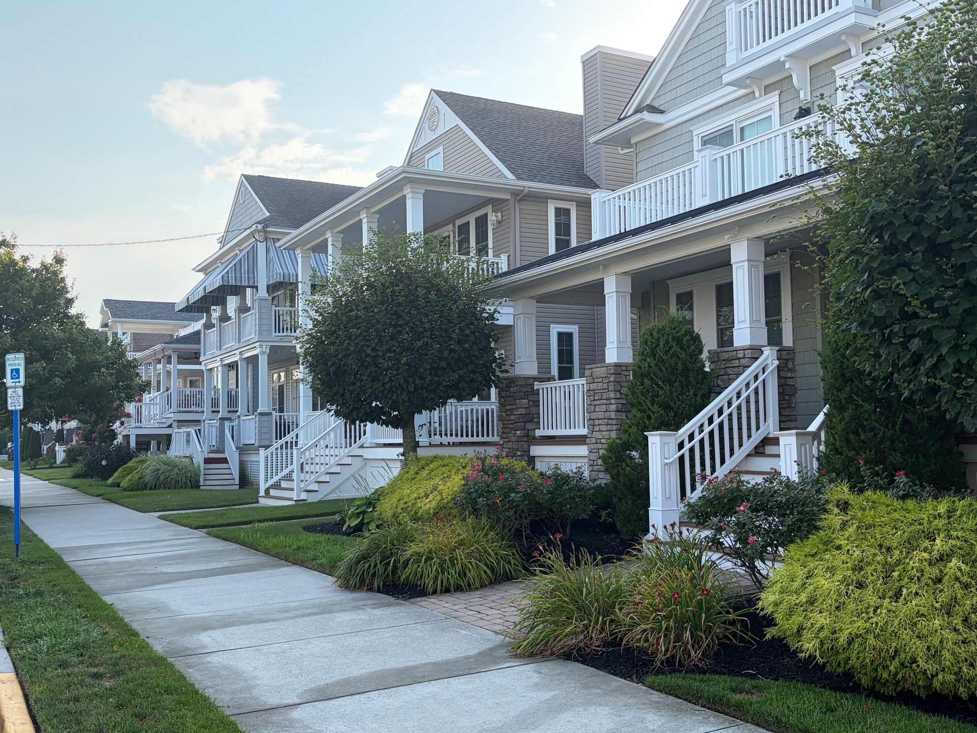 Row of white Victorian-style houses with front porches along a quiet sidewalk, surrounded by trimmed shrubs.